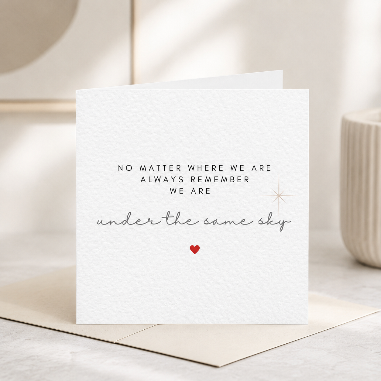 Modern memorial greeting card on white hammered cardstock with a kraft envelope. Features the quote "No Matter Where We Are, Always Remember We Are Under the Same Sky" in elegant typography with a subtle star and small red heart.