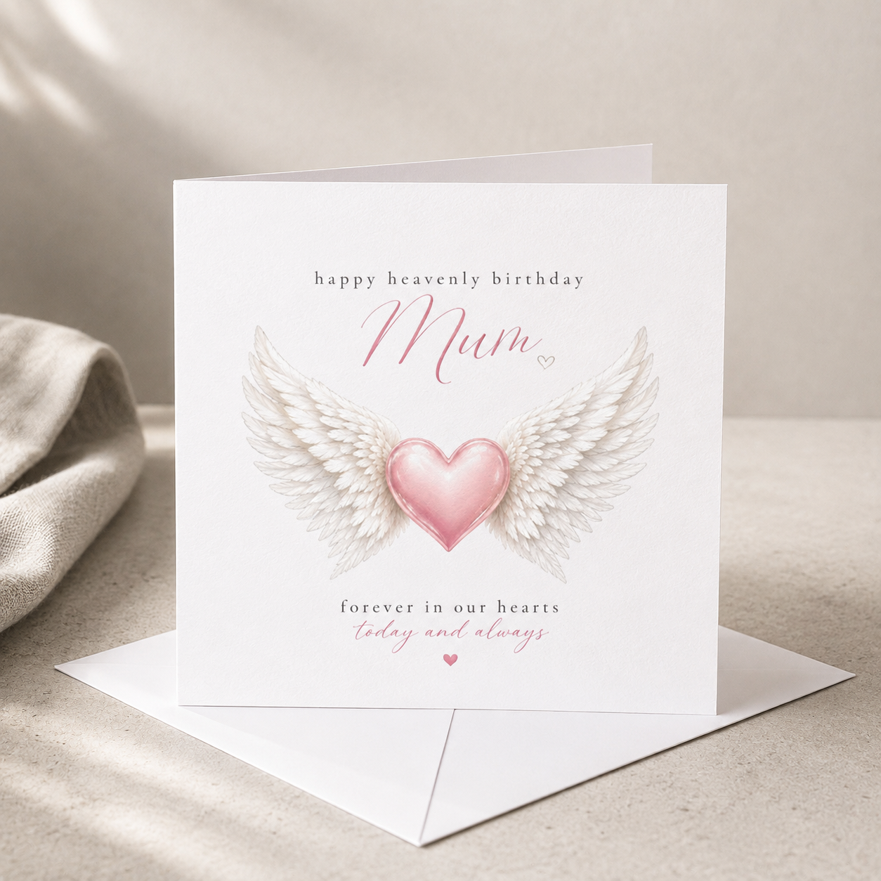 Happy Heavenly Birthday memorial card featuring angel wings and a pink heart, displayed on white hammered cardstock with a white envelope.