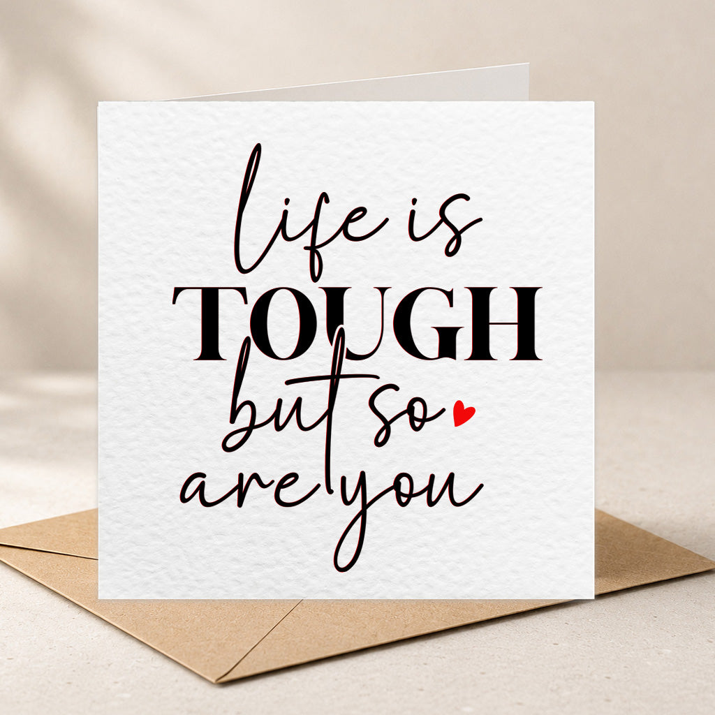 A square white greeting card with textured paper and black typographic lettering that reads 'life is TOUGH but so are you,' with a small red heart. A brown kraft envelope sits behind the card on a light beige surface.