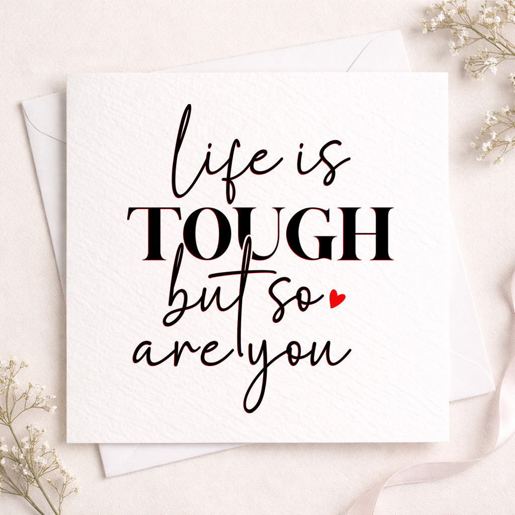 A square white greeting card with the phrase 'life is TOUGH but so are you' in black script and block lettering, with a small red heart accent, shown with white envelopes on a light blush background with delicate floral props.
