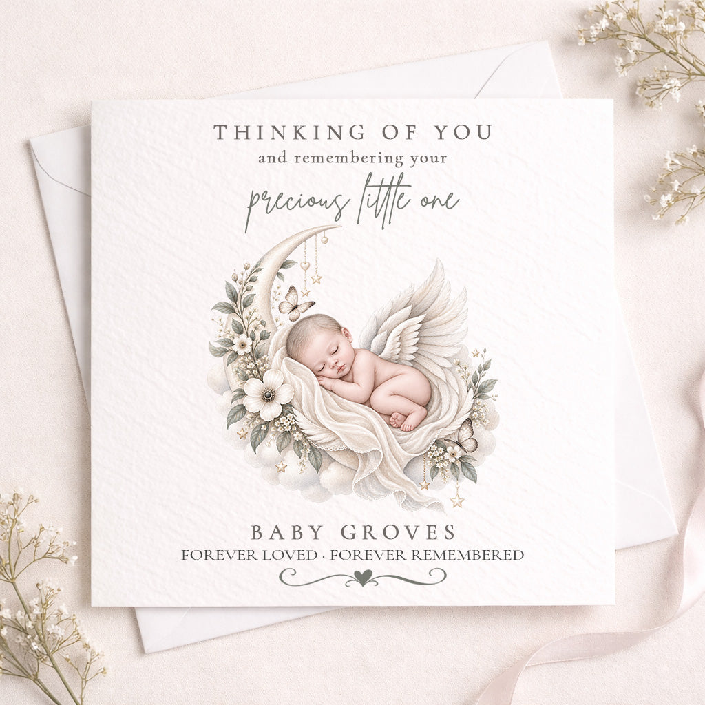 A square white memorial card with a soft illustration of a sleeping baby angel surrounded by flowers and a crescent moon, placed on top of a white envelope with delicate dried flowers around it. The card reads, 'THINKING OF YOU and remembering your precious little one' and 'BABY GROVES FOREVER LOVED - FOREVER REMEMBERED'.