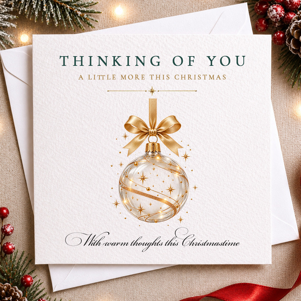 A square Christmas greeting card with a white textured cover, featuring a gold ornament illustration and the text 'THINKING OF YOU A LITTLE MORE THIS CHRISTMAS' and 'With warm thoughts this Christmasine.' The card is layered over white envelopes and surrounded by holiday decorations including pine branches, red berries, and a red ribbon on a warm beige surface.