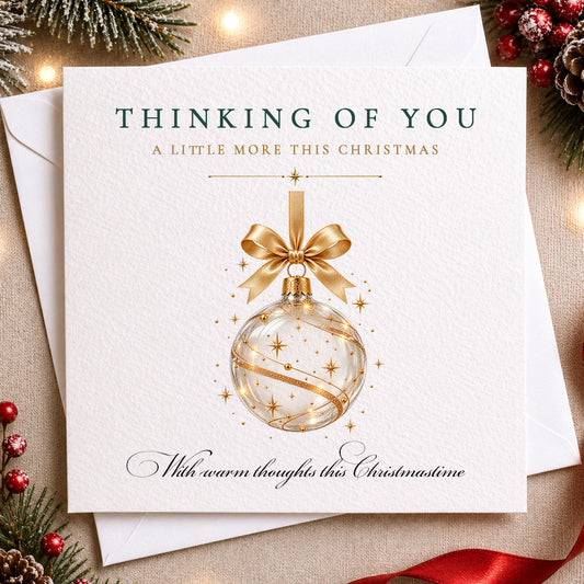 A square Christmas greeting card with a white textured cover, featuring a gold ornament illustration and the text 'THINKING OF YOU A LITTLE MORE THIS CHRISTMAS' and 'With warm thoughts this Christmasine.' The card is layered over white envelopes and surrounded by holiday decorations including pine branches, red berries, and a red ribbon on a warm beige surface.