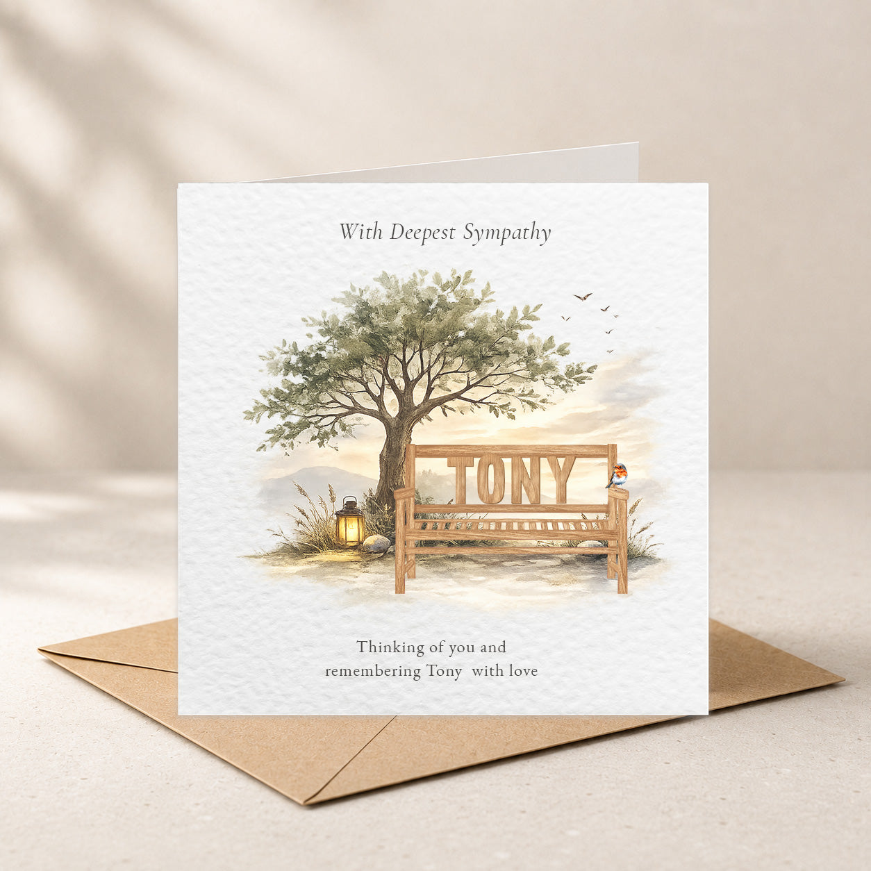 A sympathy greeting card with a watercolor illustration of a tree, a wooden bench, and the name 'TONY' on the bench, shown with a brown envelope on a light beige surface.