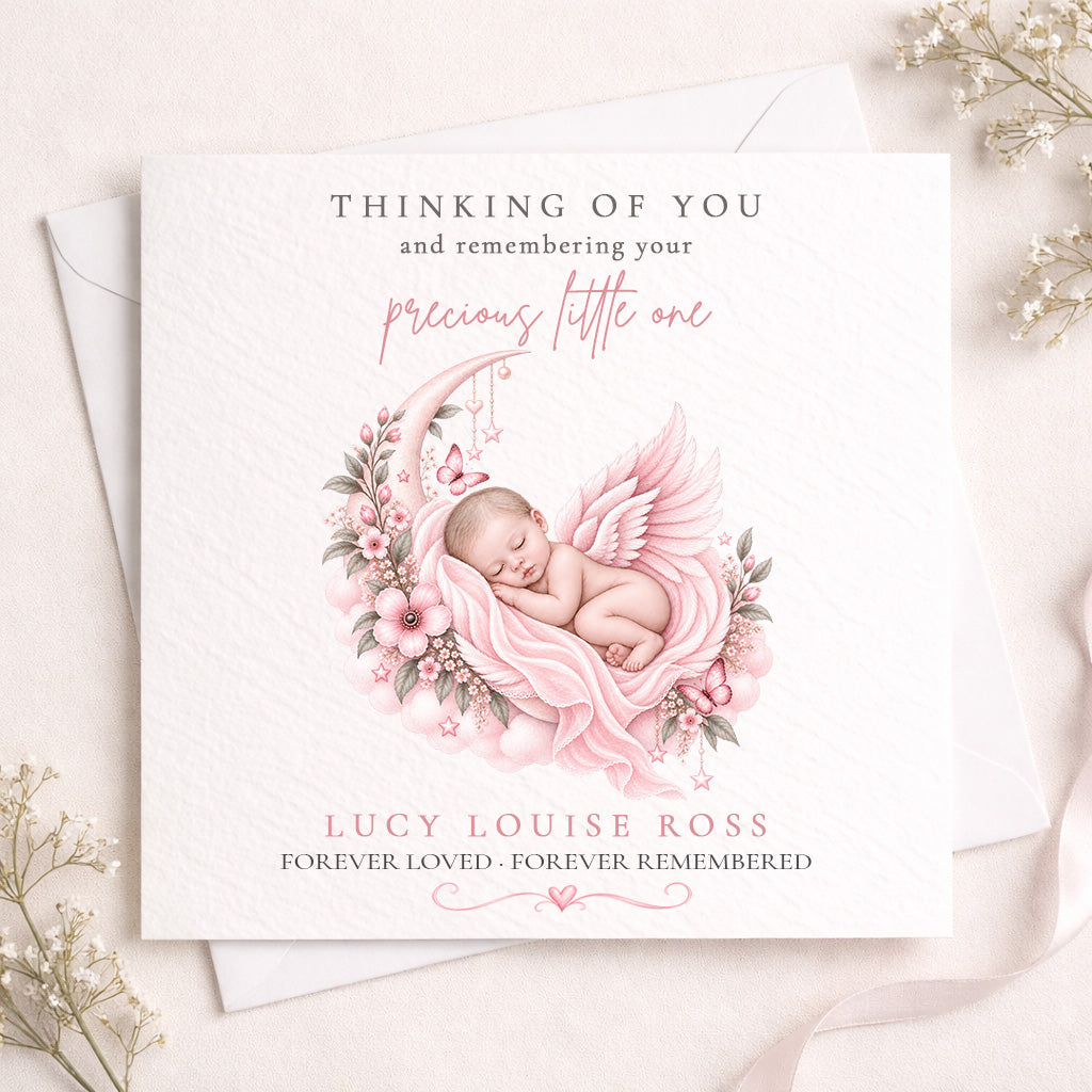 A square white memorial card with a soft pink illustration of a sleeping angel surrounded by flowers, shown on top of a white envelope. The card reads, 'THINKING OF YOU and remembering your precious little one' and 'LUCY LOUISE ROSS FOREVER LOVED - FOREVER REMEMBERED.