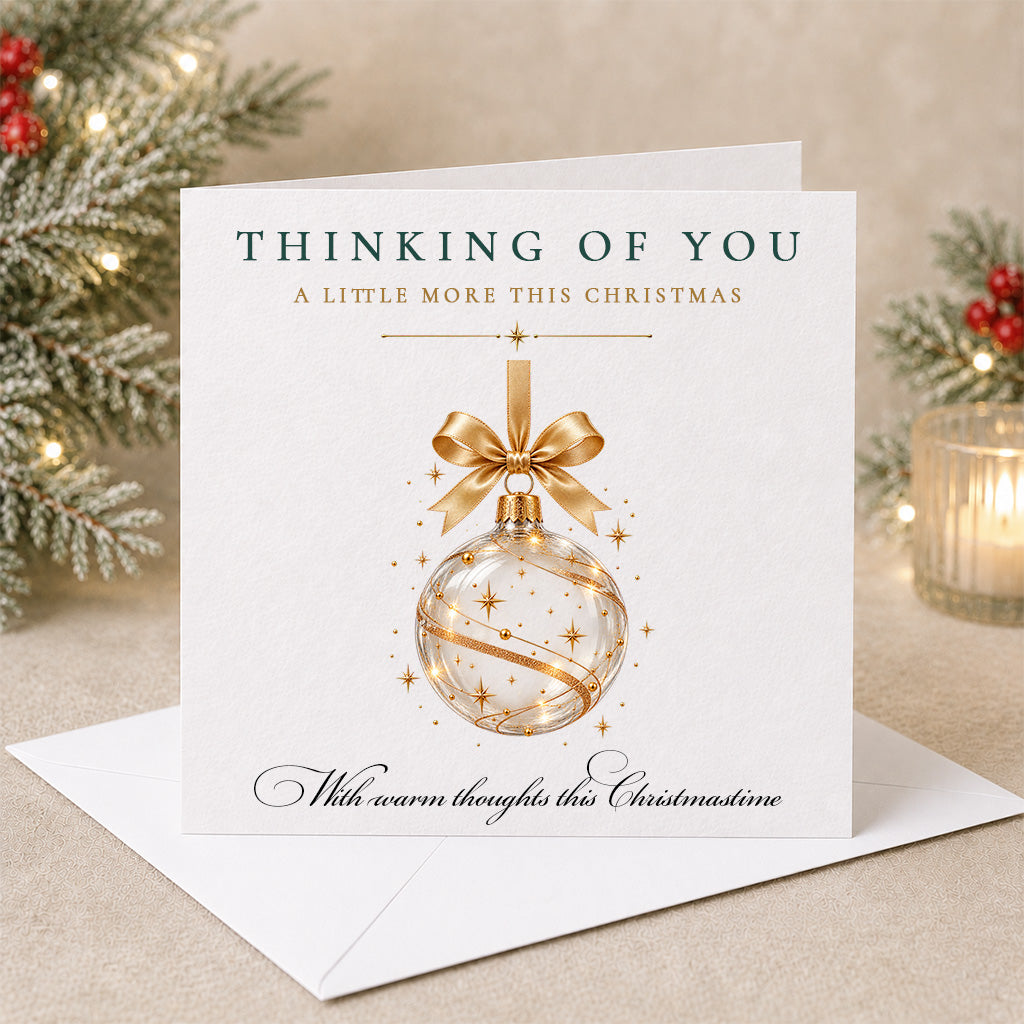 A square Christmas greeting card with a white envelope, featuring a gold ornament illustration and the text 'THINKING OF YOU A LITTLE MORE THIS CHRISTMAS' and 'With warm thoughts this Christmasine.' The card is styled on a beige surface with holiday greenery and a lit candle in the background.