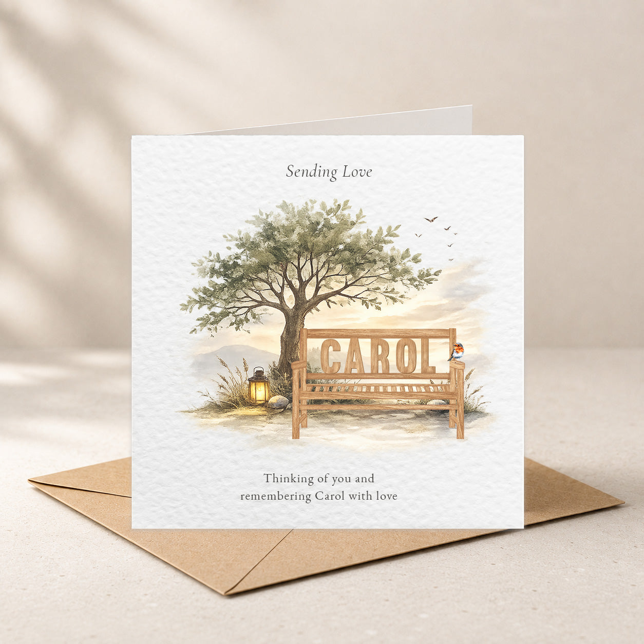 A square greeting card with a watercolor-style illustration of a tree, a wooden bench, and the name 'Carol' on the bench, shown standing on top of a brown envelope against a soft beige background.