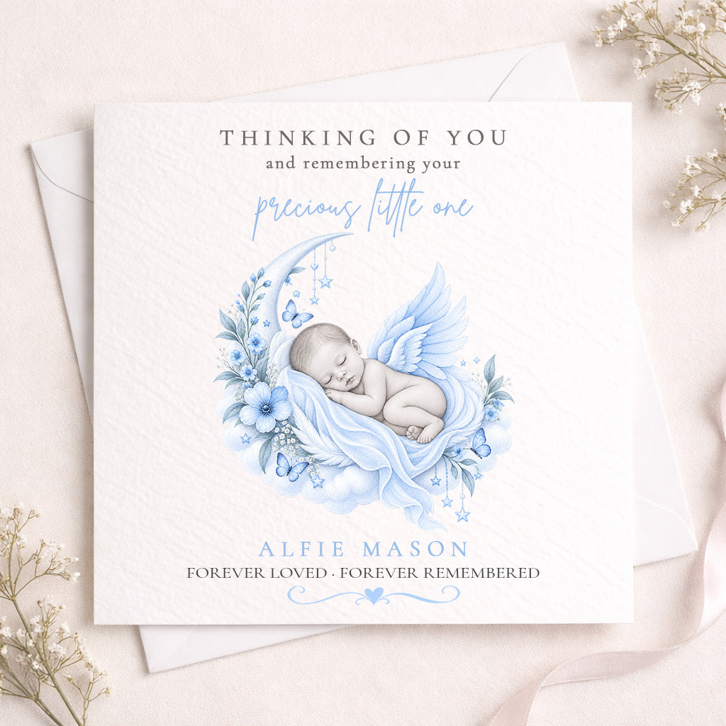 A square white memorial card with a soft blue watercolor illustration of a sleeping angel baby, layered over a white envelope. The card features personalized text reading 'THINKING OF YOU and remembering your precious little one' and 'ALFIE MASON FOREVER LOVED - FOREVER REMEMBERED'.