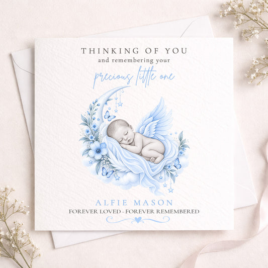 A square white memorial card with a soft blue watercolor illustration of a sleeping angel baby, layered over a white envelope. The card features personalized text reading 'THINKING OF YOU and remembering your precious little one' and 'ALFIE MASON FOREVER LOVED - FOREVER REMEMBERED'.