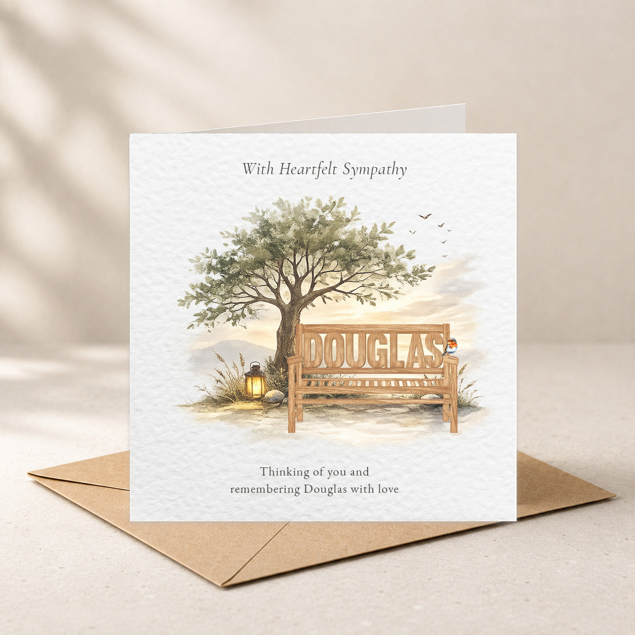 A sympathy greeting card with a watercolor illustration of a tree, bench, and small lantern, with the name DOUGLAS on the bench. The card is shown standing upright on a brown envelope against a soft beige background.