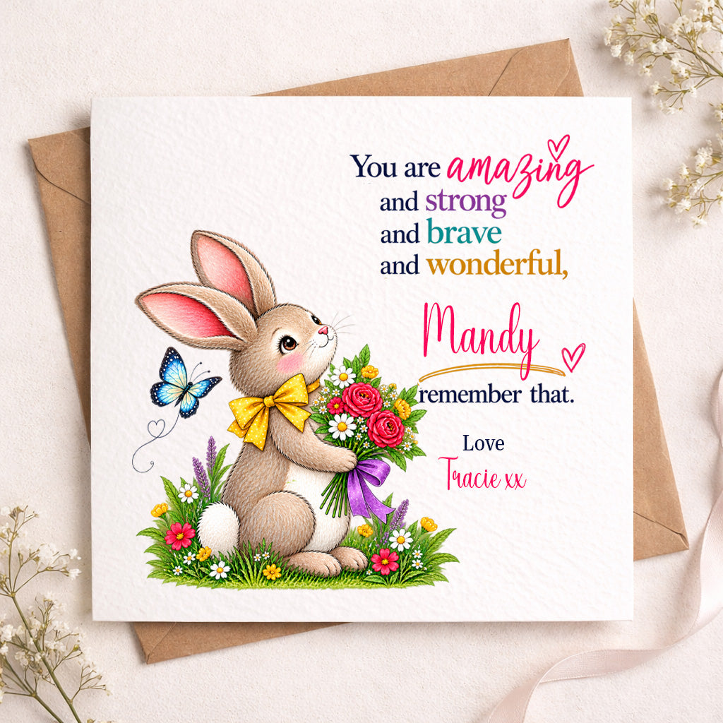 A square greeting card featuring an illustrated brown bunny holding a bouquet of red, pink, and white flowers with a yellow bow, next to a butterfly. The card has colorful text reading, 'You are amazing and strong and brave and wonderful, Mandy remember that.