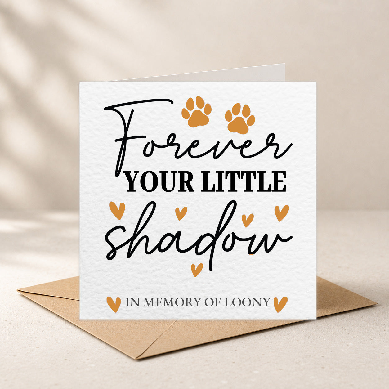 A square white memorial card with the words 'Forever YOUR LITTLE shadow IN MEMORY OF LOONY' in black script and block lettering, decorated with two orange paw prints and small orange hearts. The card is shown standing upright on a brown envelope against a warm beige background with soft leaf shadows.
