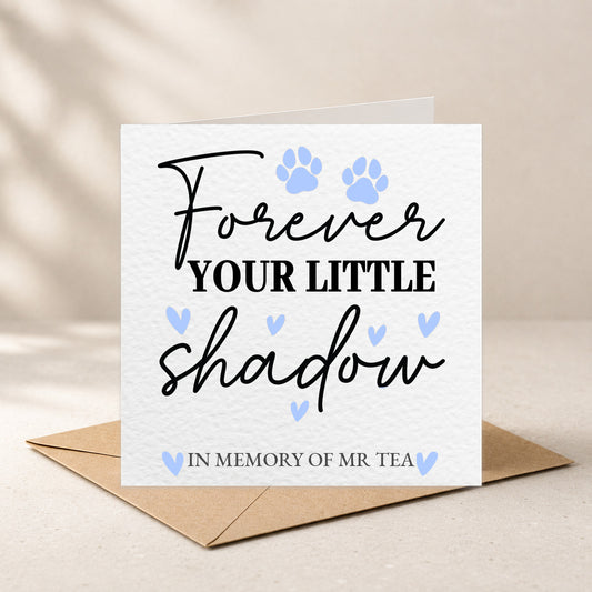 A square white memorial card with the text 'Forever YOUR LITTLE shadow IN MEMORY OF MR TEA' in black script and block lettering, decorated with two light blue paw prints and small blue hearts. The card is shown standing upright on a brown envelope against a warm beige background.