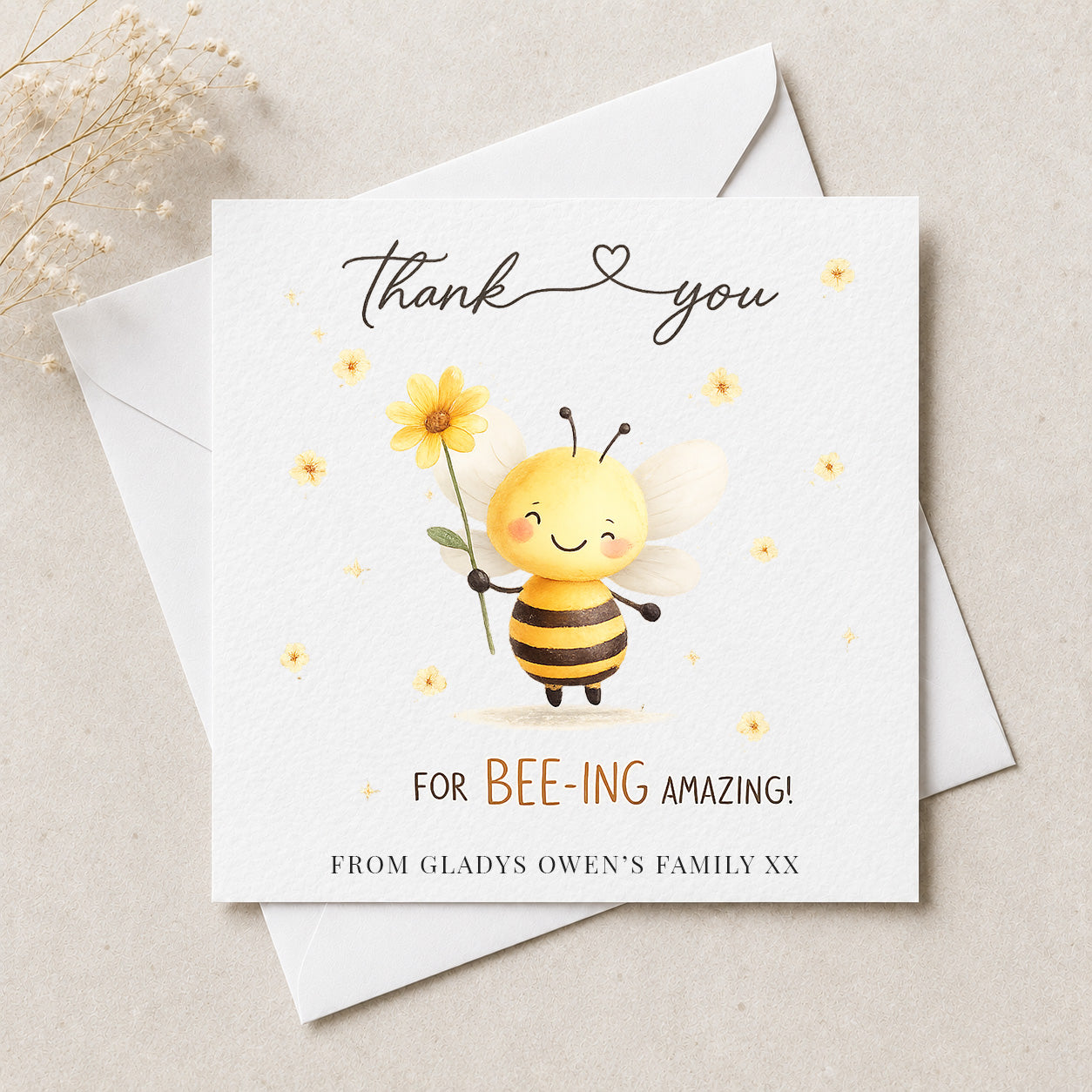 A square white thank-you card with a cute cartoon bee holding a yellow flower, layered over a white envelope on a light beige surface. The card reads 'thank you' with a heart between the words, 'FOR BEE-ING AMAZING!' in black and orange text, and 'FROM GLADYS OWEN'S FAMILY XX' at the bottom.