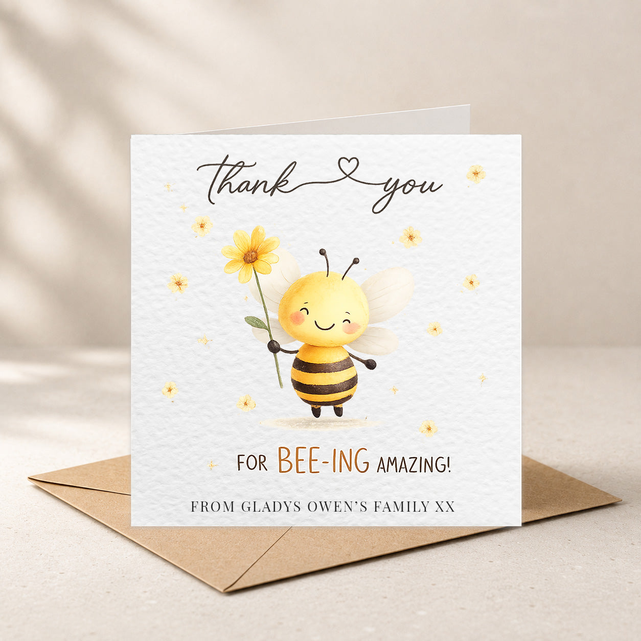 A square white greeting card with a cute cartoon bee holding a yellow flower, shown standing on top of a brown envelope against a warm beige background. The card reads 'thank you' and 'FOR BEE-ING AMAZING!' with a personalized line at the bottom.
