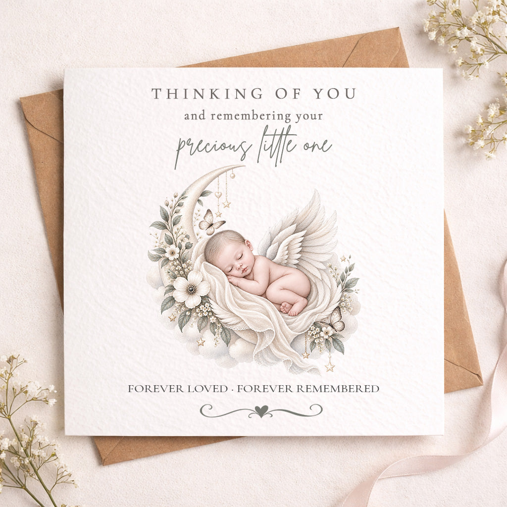 A square white memorial card with a printed illustration of a sleeping baby angel surrounded by flowers and a crescent moon, placed on top of a brown kraft envelope. The card reads, 'THINKING OF YOU and remembering your precious little one' and 'FOREVER LOVED - FOREVER REMEMBERED.