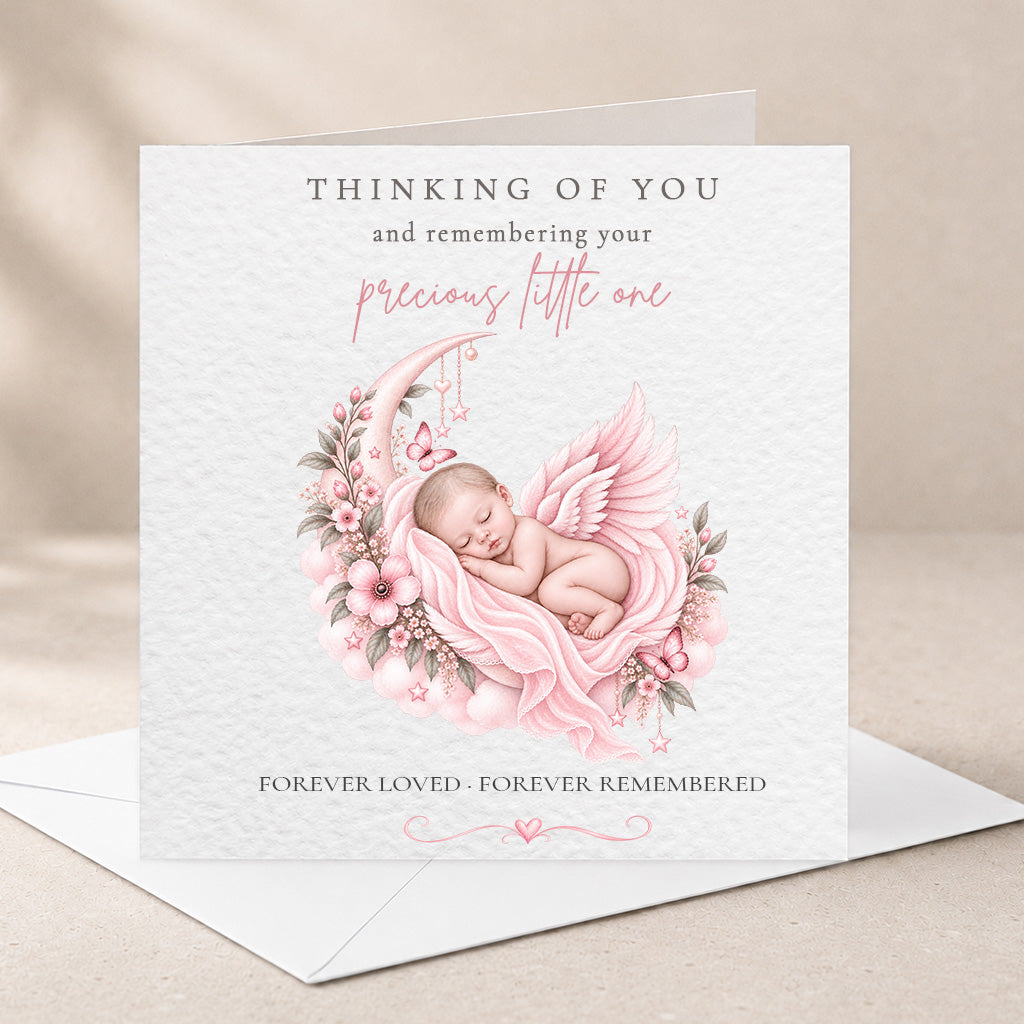 A square white memorial card with a soft pink illustration of a sleeping baby angel surrounded by flowers, shown standing upright on a white envelope against a warm beige background.