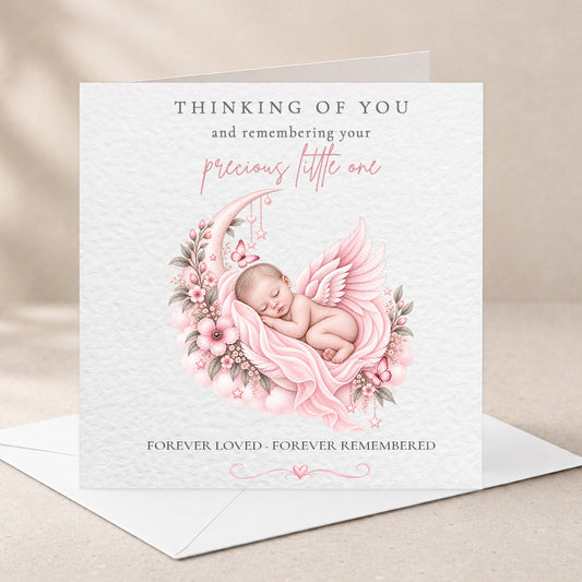 A square white memorial card with a soft pink illustration of a sleeping baby angel surrounded by flowers, shown standing upright on a white envelope against a warm beige background.