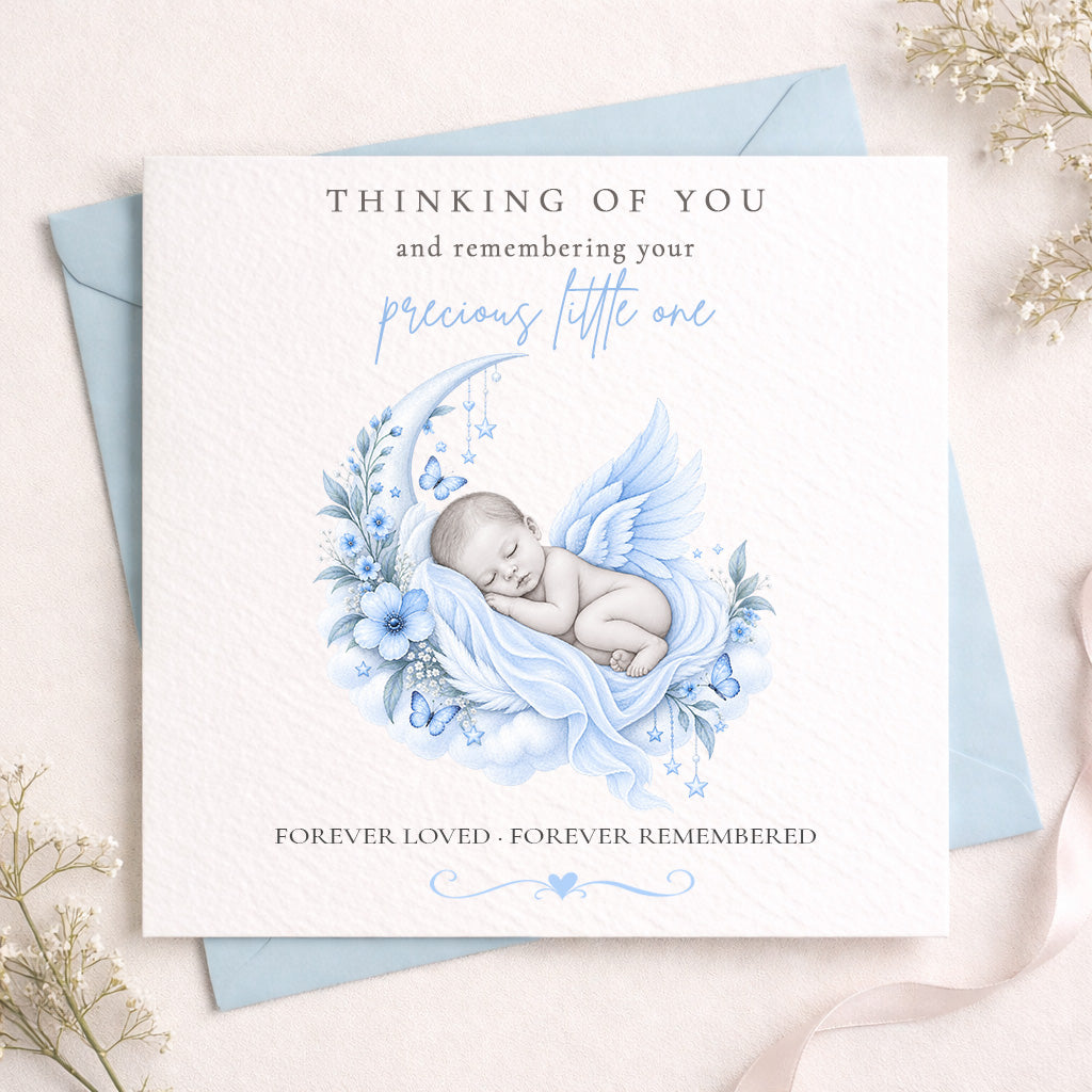 A square white memorial card with a light blue envelope behind it, styled on a pale pink surface with dried flowers. The card reads 'THINKING OF YOU and remembering your precious little one' and 'FOREVER LOVED - FOREVER REMEMBERED,' with a soft illustration of a sleeping baby in a blue crescent moon and floral wreath.