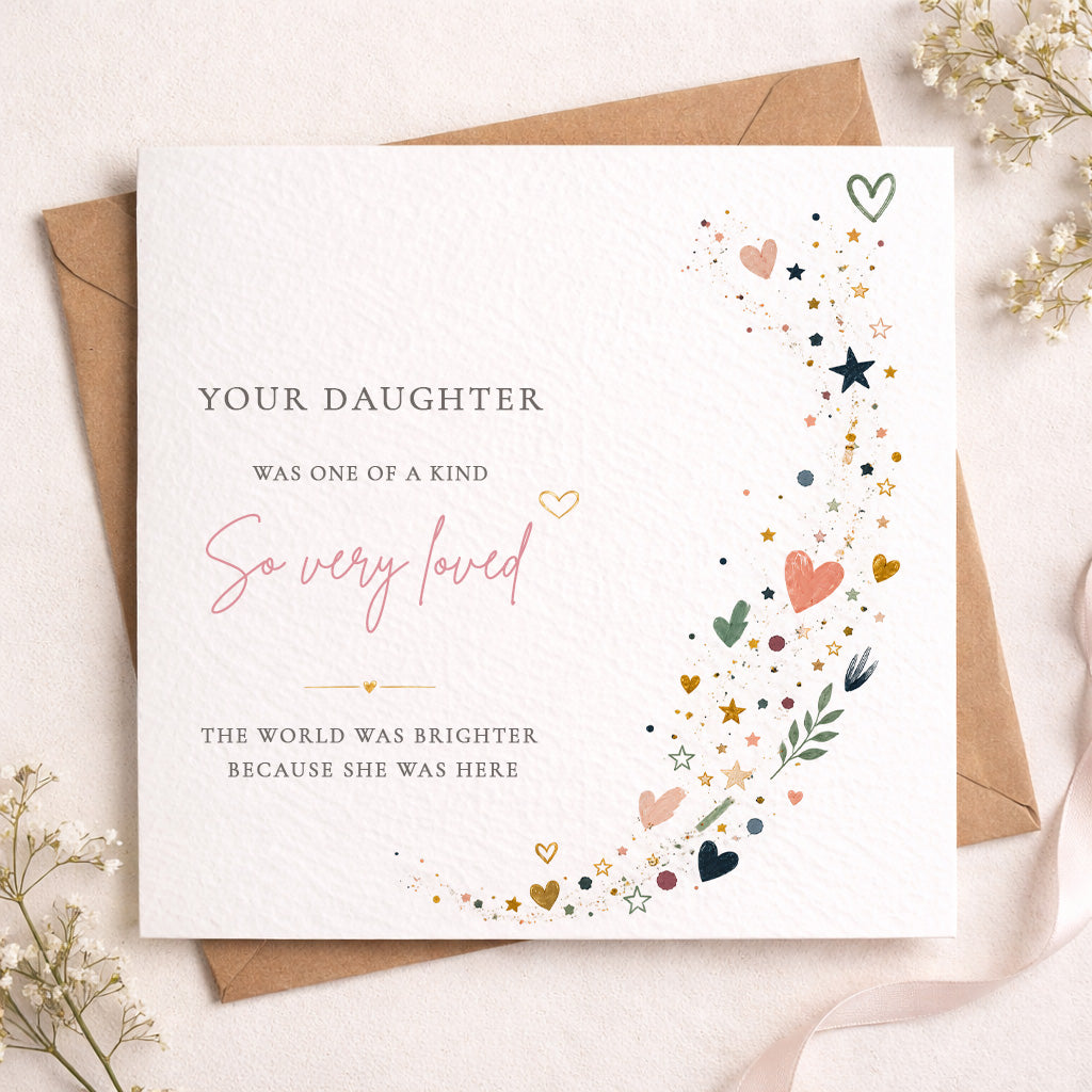 A square white memorial card with printed text and delicate multicolor heart, star, and leaf illustrations, shown with a brown envelope on a soft beige surface with dried flowers.