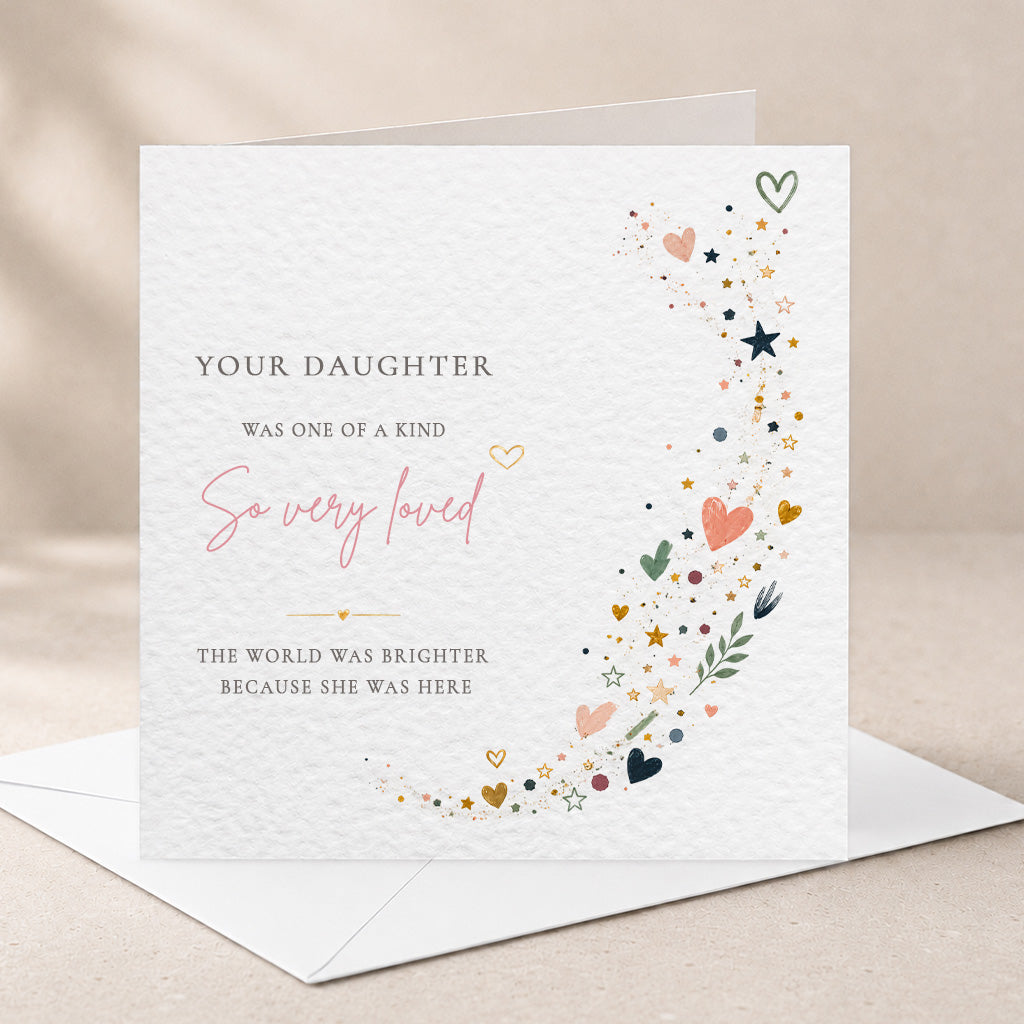 A square white greeting card with a watercolor-style decorative border of stars, hearts, and leaves along the right side, placed on top of a white envelope against a warm beige background. The card reads, 'YOUR DAUGHTER WAS ONE OF A KIND So very loved THE WORLD WAS BRIGHTER BECAUSE SHE WAS HERE.