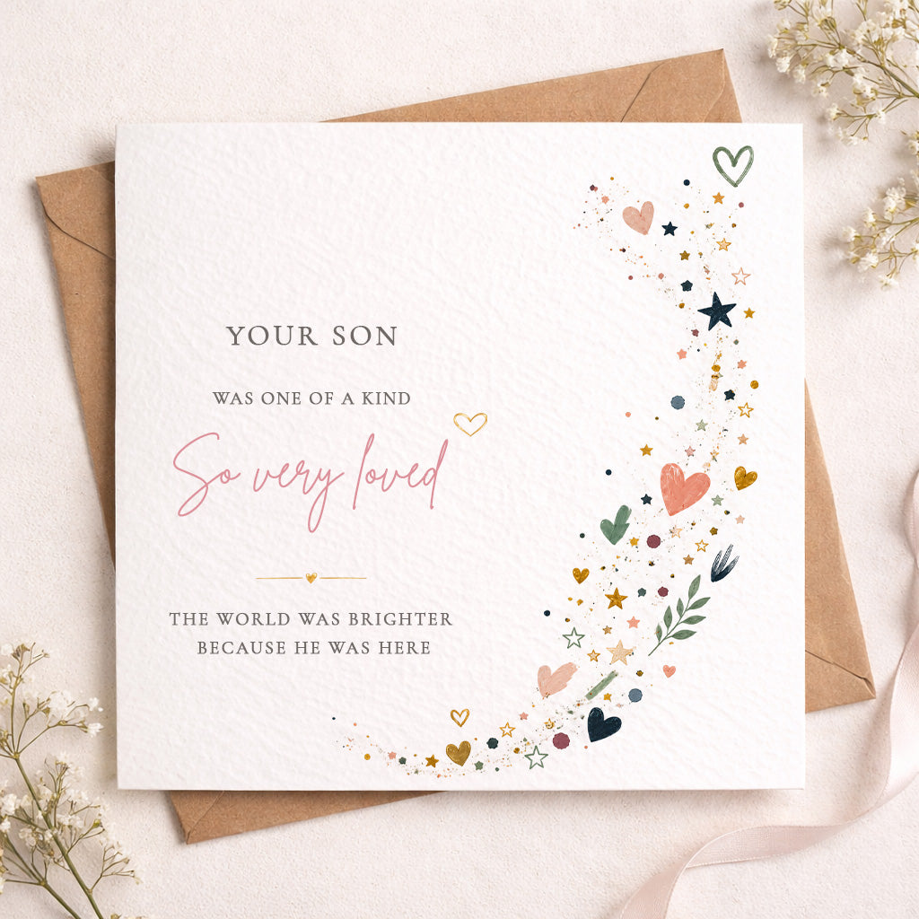 A square white memorial or remembrance card with printed text and delicate multicolor heart, star, and leaf illustrations, shown with a brown envelope on a light fabric background with small dried flowers.