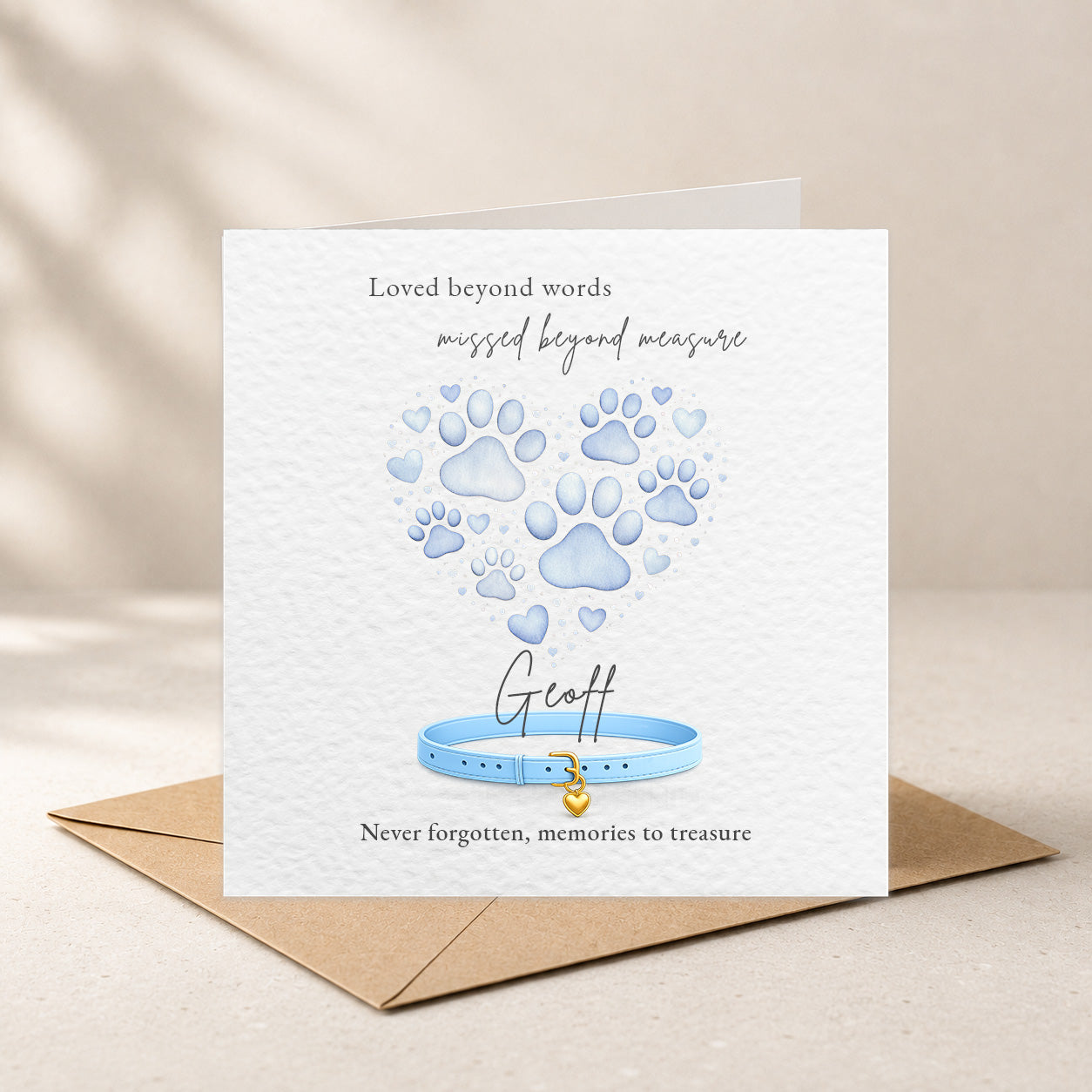 A square white memorial card with a kraft envelope behind it, featuring blue paw prints and a blue collar illustration with the text 'Loved beyond words missed beyond measure' and 'Never forgotten, memories to treasure.' The card is shown standing upright on a light beige surface with soft shadow overlays.