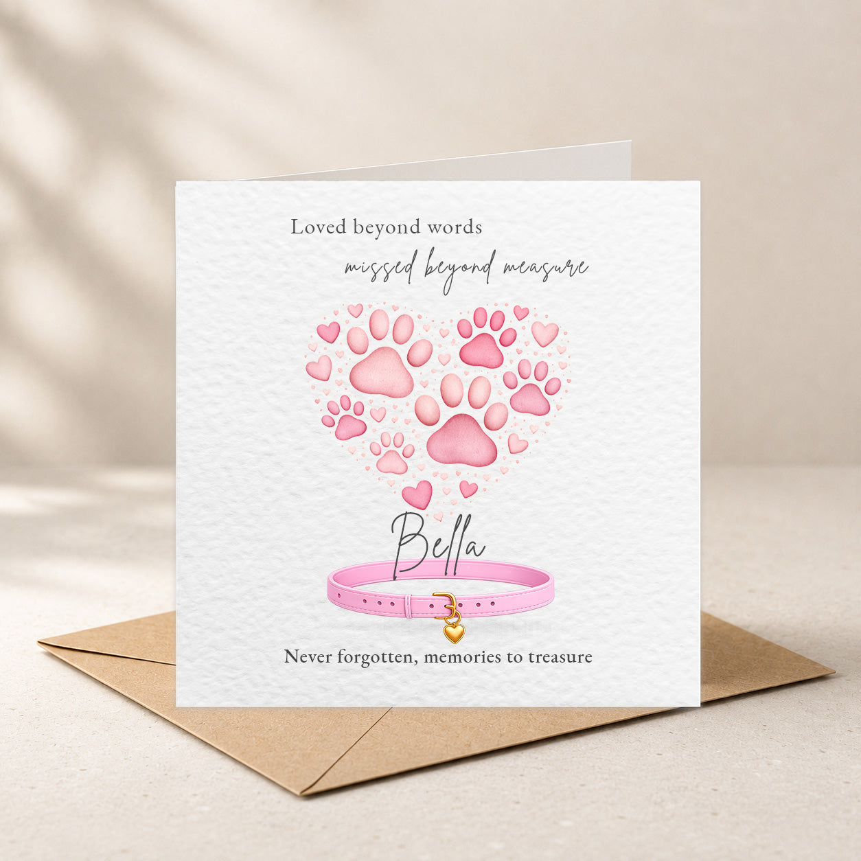 A square white memorial card with pink paw prints forming a heart, a pink collar illustration, and the name 'Bella' in script, shown standing upright on a brown envelope against a softly lit beige background.