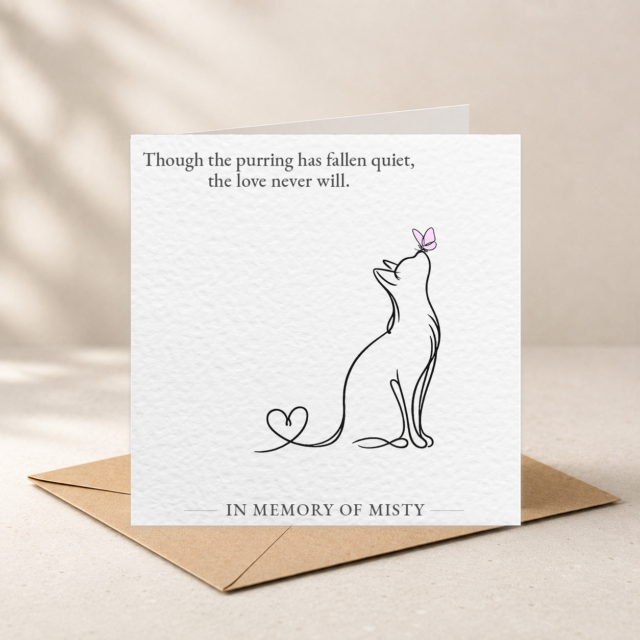 A square white memorial card with printed text and a simple line drawing of a cat, shown standing upright on a brown kraft envelope against a warm beige background.