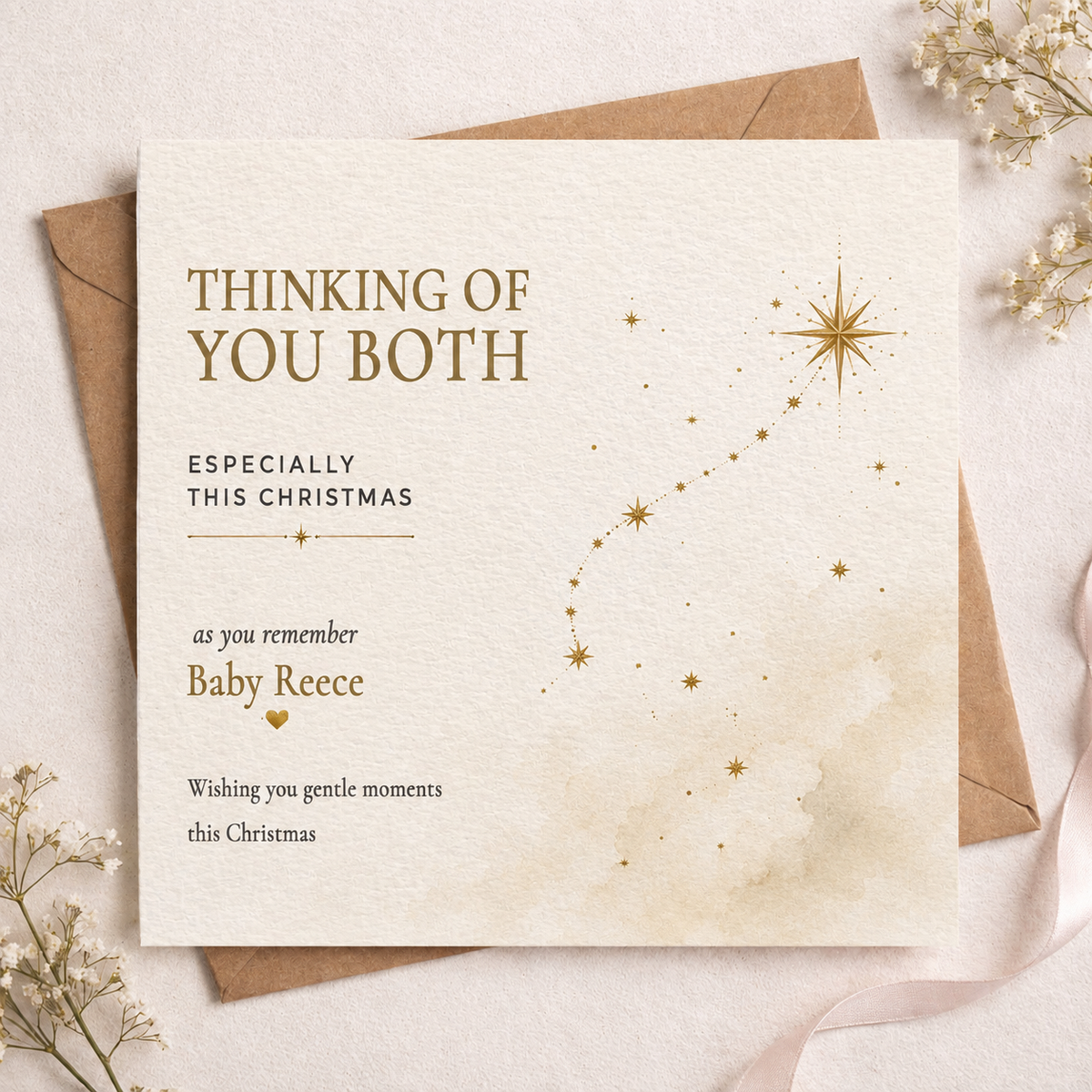 A square Christmas greeting card with a warm cream background and gold text reading 'THINKING OF YOU BOTH ESPECIALLY THIS CHRISTMAS' and 'as you remember Baby Reece,' styled on top of brown envelopes with dried flowers and a ribbon.