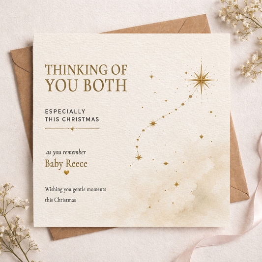 A square Christmas greeting card with a warm cream background and gold text reading 'THINKING OF YOU BOTH ESPECIALLY THIS CHRISTMAS' and 'as you remember Baby Reece,' styled on top of brown envelopes with dried flowers and a ribbon.