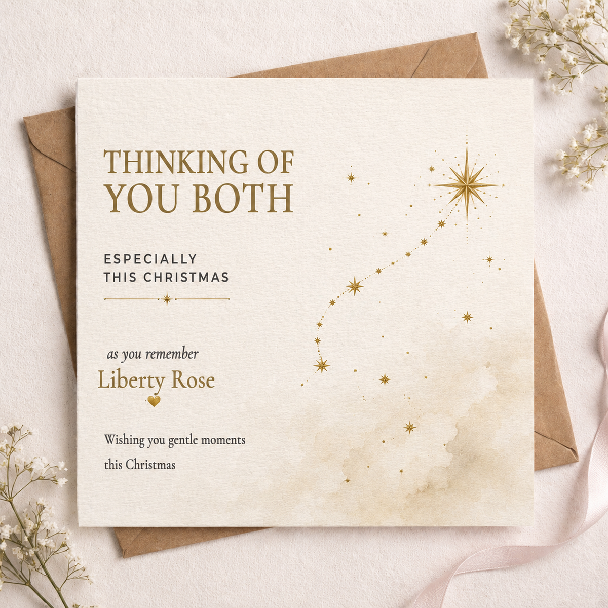A square Christmas greeting card with a textured cream background and elegant gold typography, styled on top of a brown envelope with delicate white flowers in the corners. The card reads 'THINKING OF YOU BOTH' and includes a gold star constellation graphic.