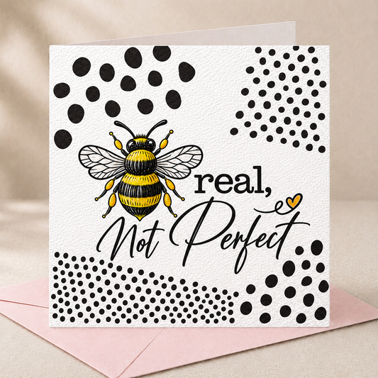 A square greeting card with a white textured background, featuring a black and yellow bumblebee illustration, black polka dot patterns, and the phrase 'real, Not Perfect' in black script and print. The card is shown standing upright on a pink envelope against a soft beige fabric background.