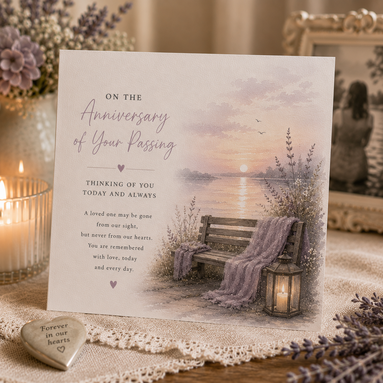 A square anniversary of your passing greeting card displayed on a tabletop with a candle, lace cloth, and floral decor in the background. The card features a soft pastel sunset over water with a bench, lavender blanket, and lantern, plus a poem of remembrance.
