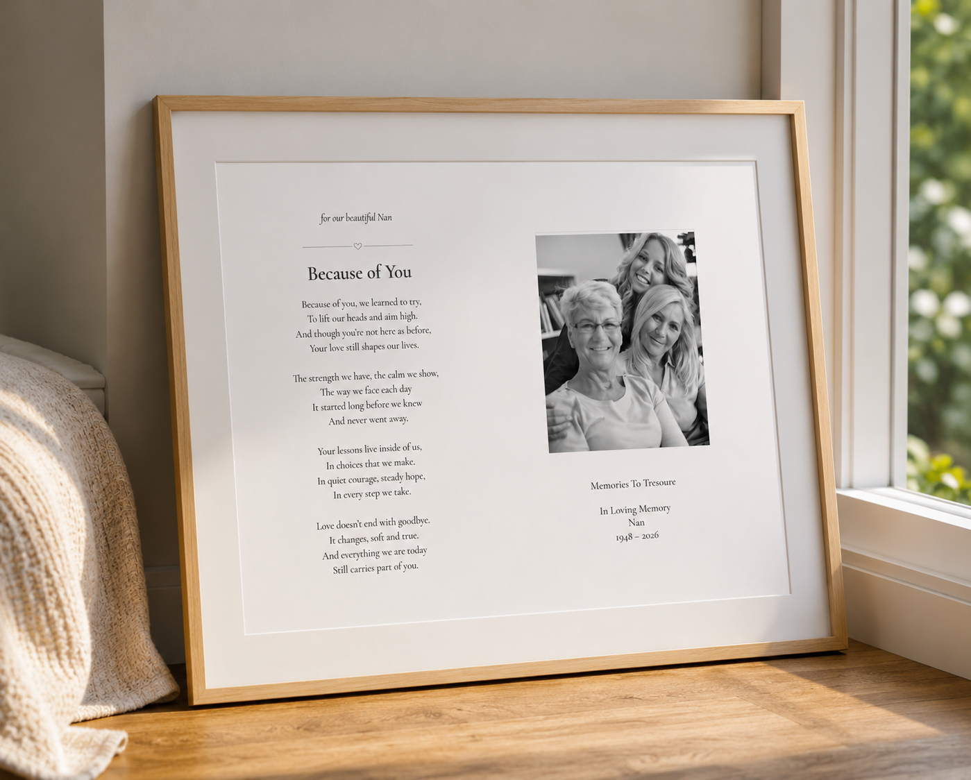 A framed personalized memorial print leaning against a wall on a wooden surface near a window. The print has a white background with a light wood frame, a poem titled 'Because of You,' a black-and-white photo of three women, and memorial text.
