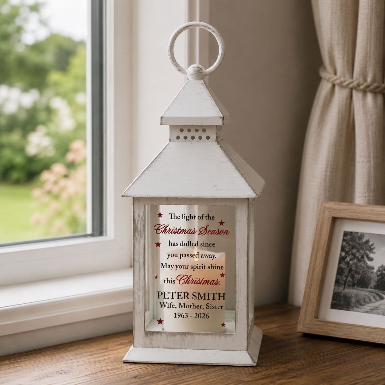 A white wooden lantern-style memorial candle holder sits on a windowsill in a home setting. The front features a printed personalized Christmas remembrance message with the name 'PETER SMITH' and dates '1963 - 2026'.