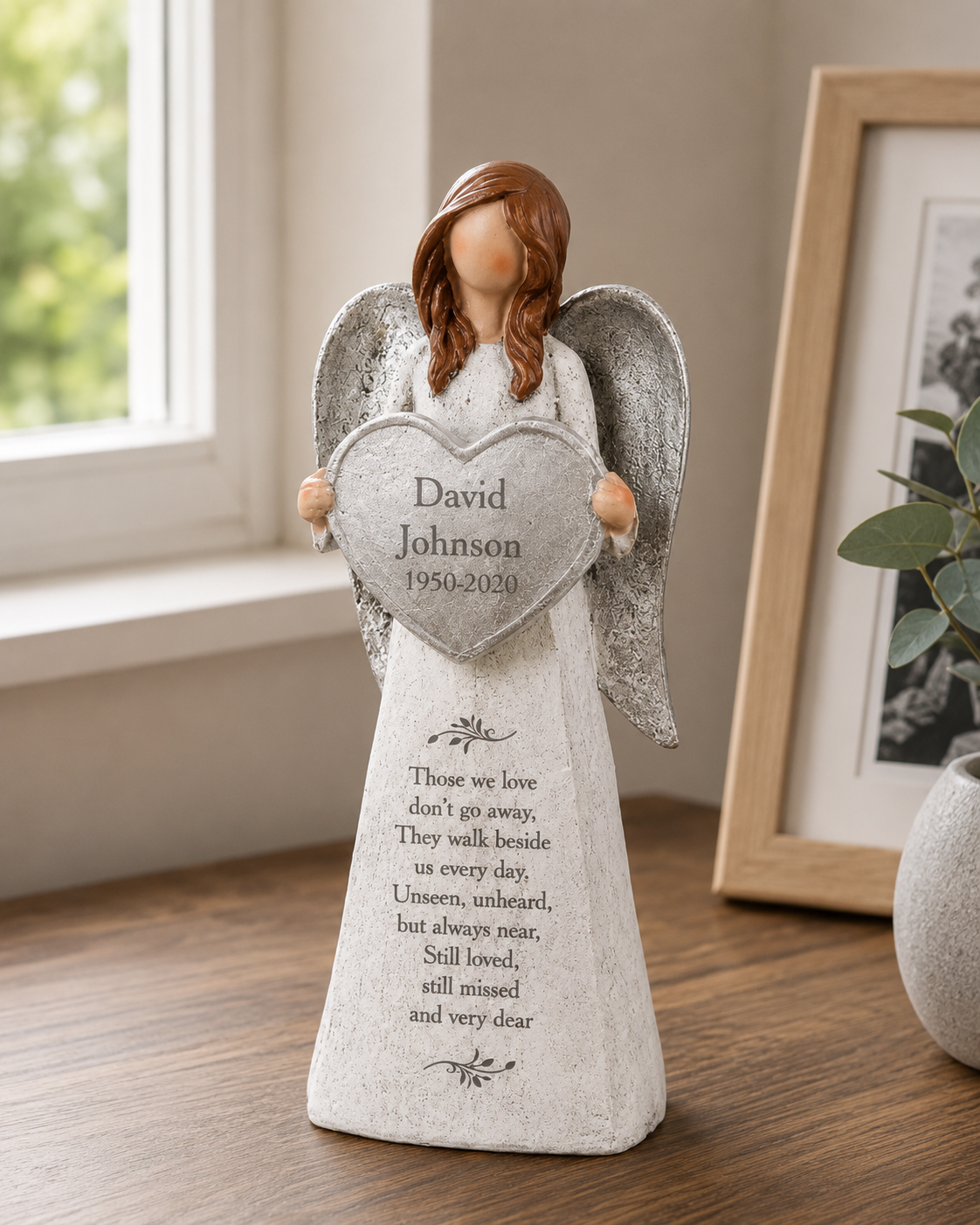 Personalized angel memorial figurine holding a heart-shaped plaque with the name 'David Johnson 1950-2020' and a poem printed on the front, displayed on a wooden surface near a window and framed picture.