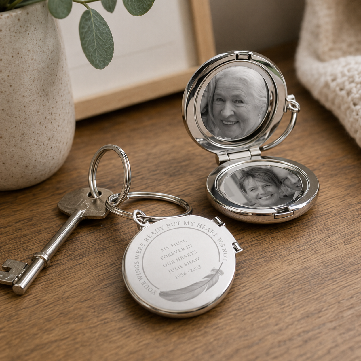 A silver keepsake photo locket keychain shown on a wooden table, with one locket open to display a printed portrait of a woman and the other closed with engraved text and a key attached.