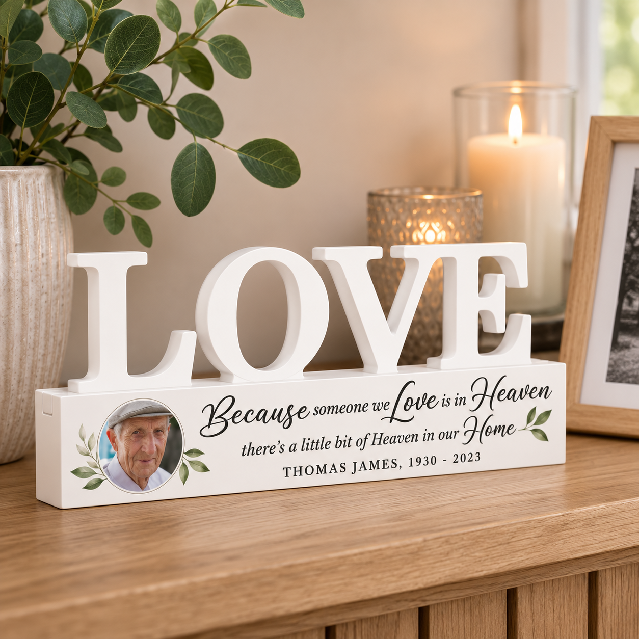 A personalized wooden 'LOVE' sign displayed on a wooden console table in a cozy home setting. The white block letters spell 'LOVE' and the base features a custom printed photo of a man, a quote, and the names 'THOMAS JAMES, 1930 - 2023'.