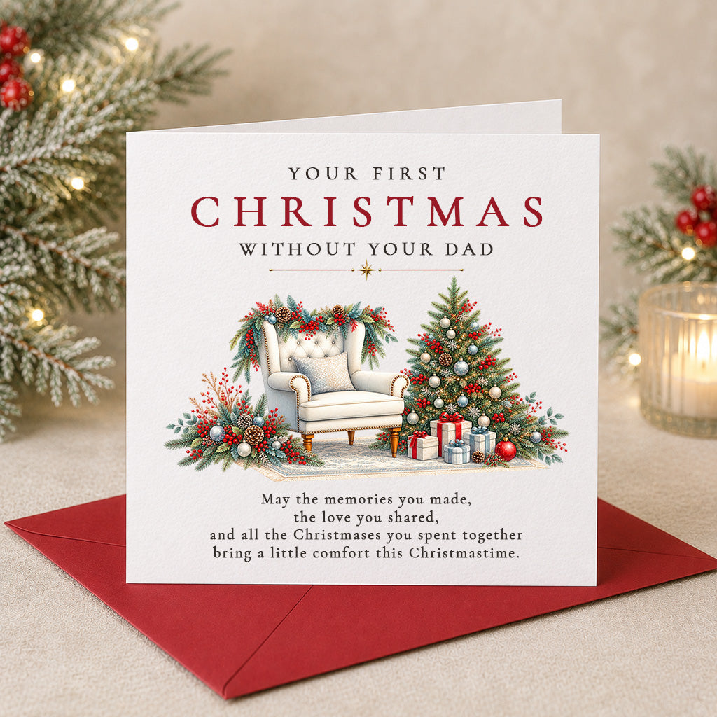 A Christmas greeting card with a red envelope, shown on a tabletop with holiday decorations in the background. The card reads 'YOUR FIRST CHRISTMAS WITHOUT YOUR DAD' and features an illustration of a decorated armchair and Christmas tree.