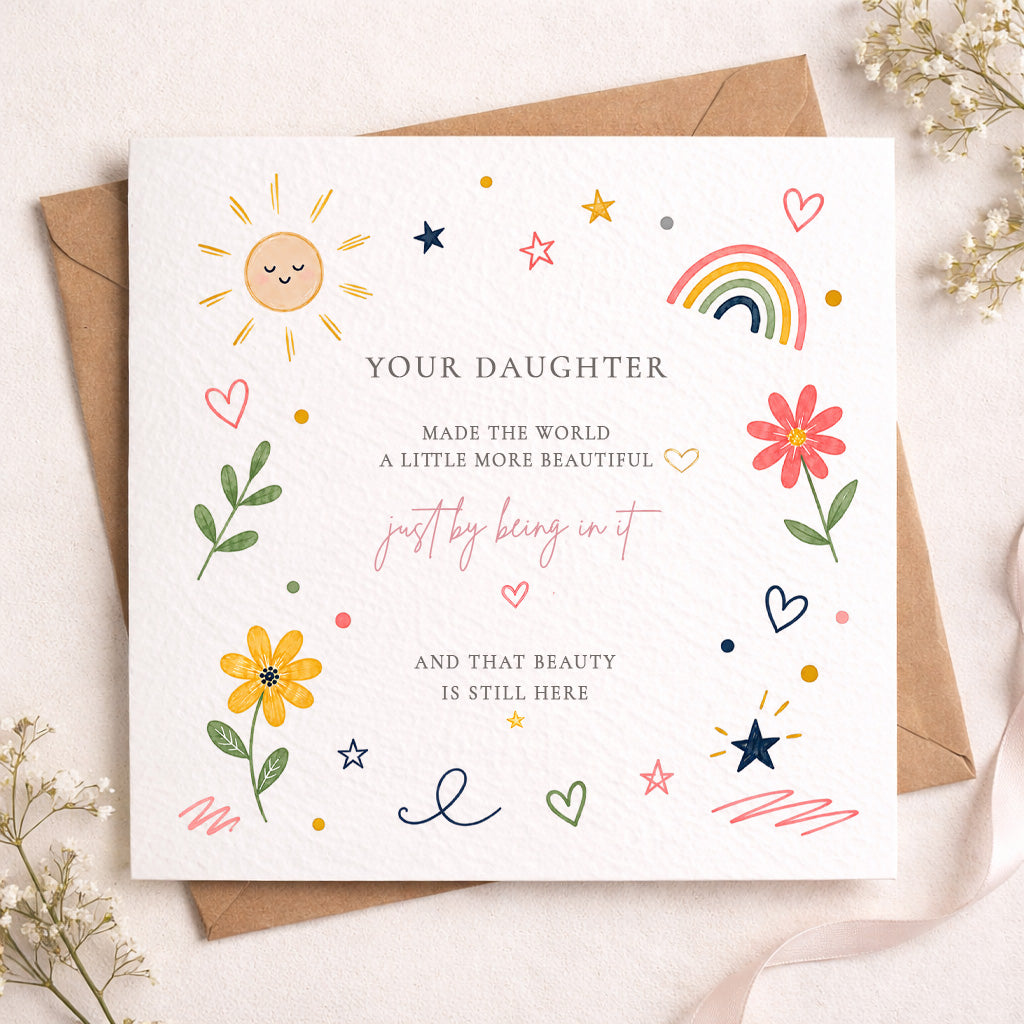 A square white greeting card with colorful hand-drawn illustrations and a personalized message for a daughter, placed on top of a brown kraft envelope with small dried flowers around it.
