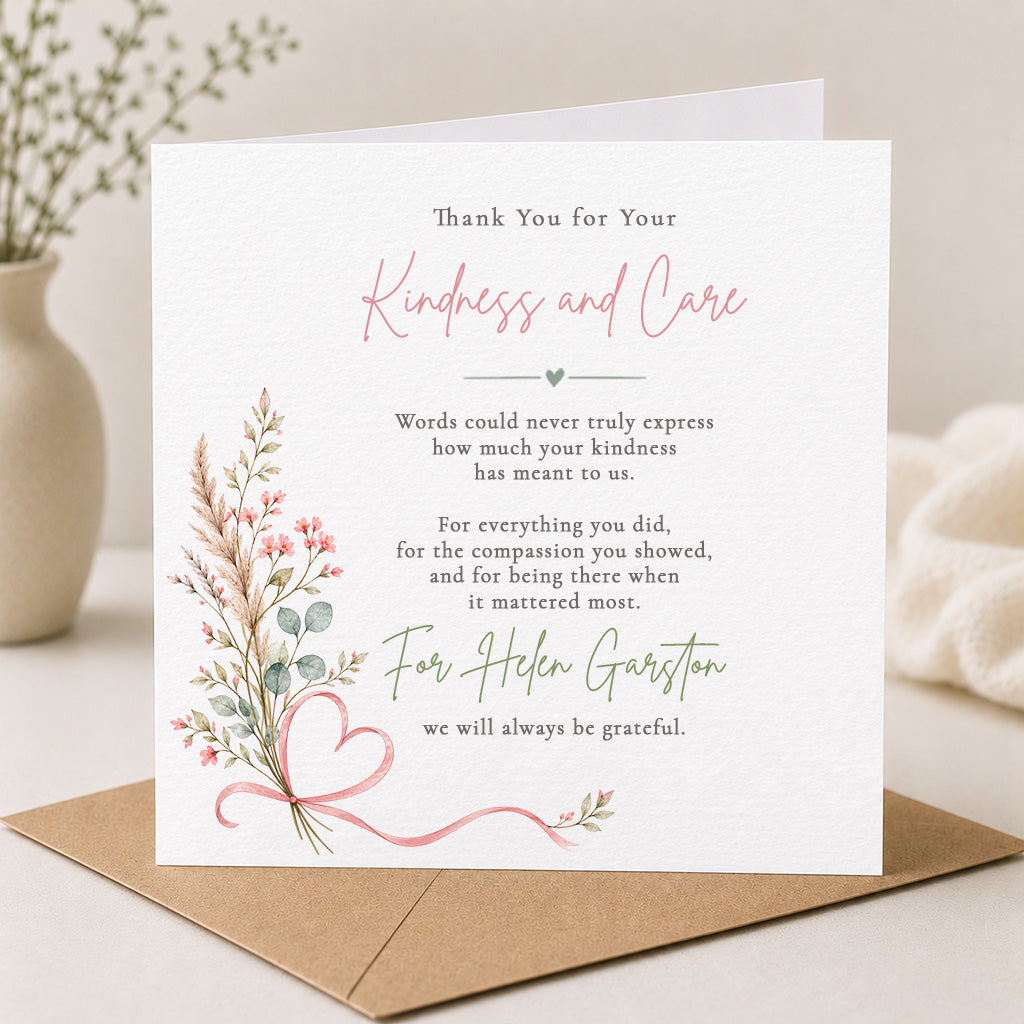 A square white sympathy or thank-you card with elegant printed text, pink and green floral accents, and a pink ribbon illustration, shown standing on a brown kraft envelope against a soft neutral background.