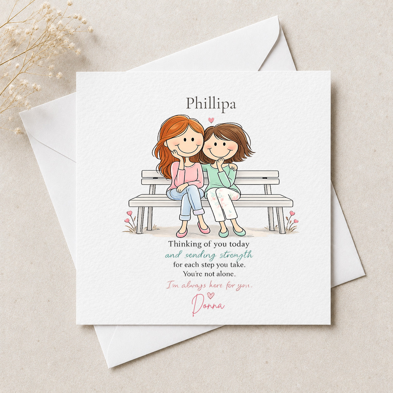 A personalized greeting card with a white textured background, featuring a cartoon illustration of two women sitting on a bench, with the name 'Phillipa' at the top and a message of support. The card is displayed with white envelopes on a light beige textured surface with dried flowers in the corner.