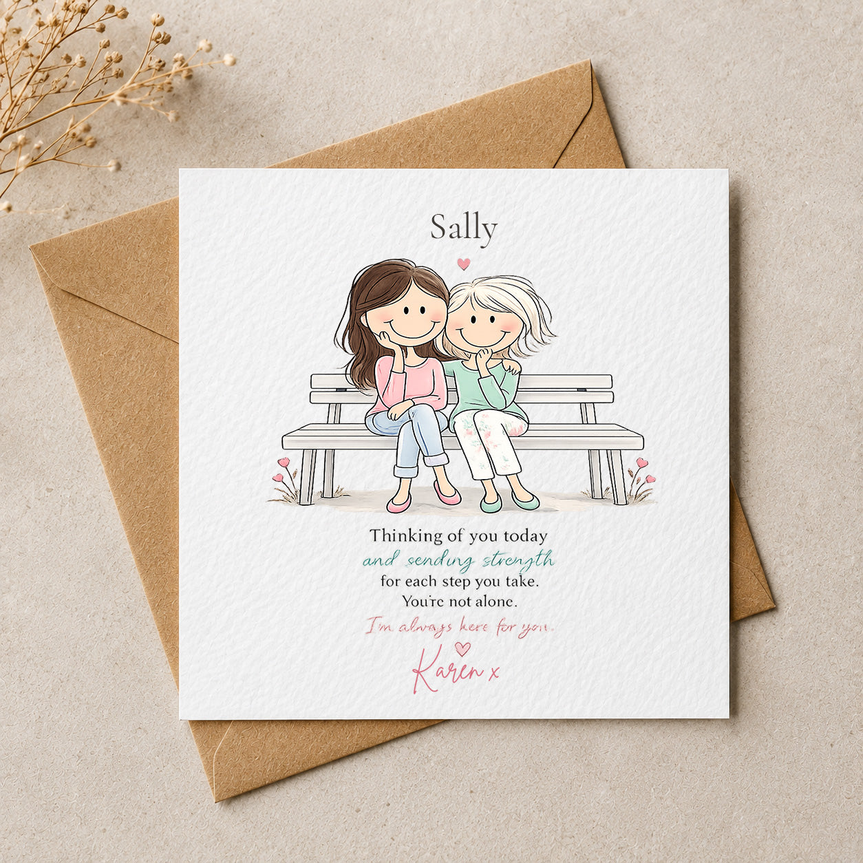A square white greeting card with a personalized illustration of two women sitting on a bench, placed on top of a brown envelope on a beige surface. The card includes the name 'Sally' at the top and a printed message, with 'Karen x' written in pink at the bottom.
