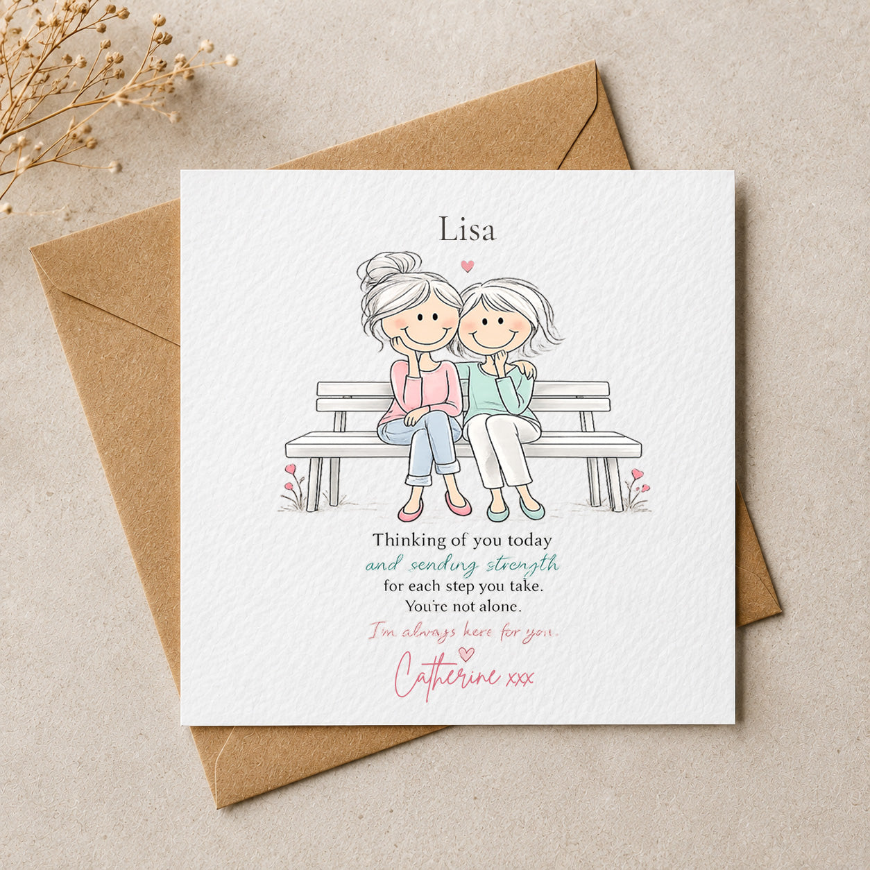 A square white memorial card with a brown kraft envelope behind it, placed on a beige surface with small dried flowers in the corner. The card features a simple illustration of two women sitting together on a bench, with the name 'Lisa' at the top and a memorial message below in black and pink text.