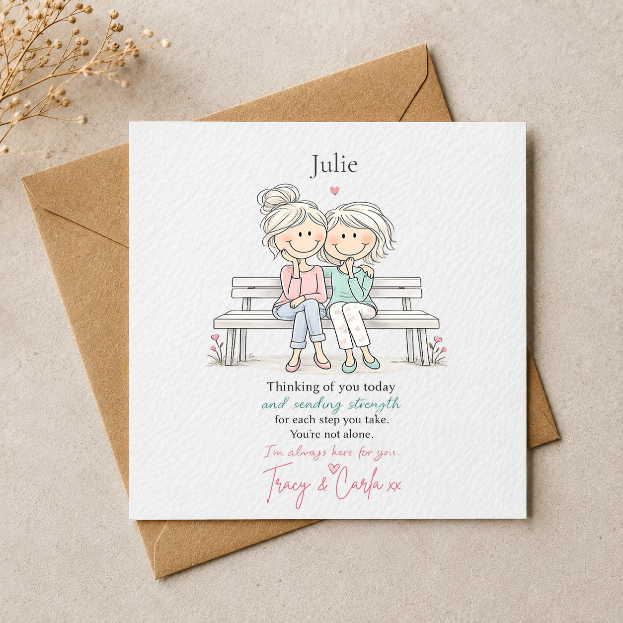 A personalized square greeting card with a brown kraft envelope, featuring an illustration of two women sitting on a bench with a printed message. The card is placed on a neutral beige surface with dried flowers in the corner.