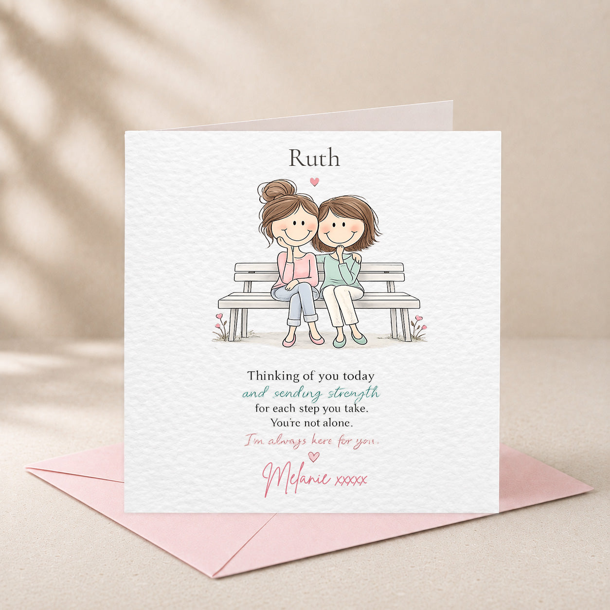 A personalized greeting card with a watercolor illustration of two women sitting on a bench, shown with a pink envelope on a beige background with soft leaf shadows.