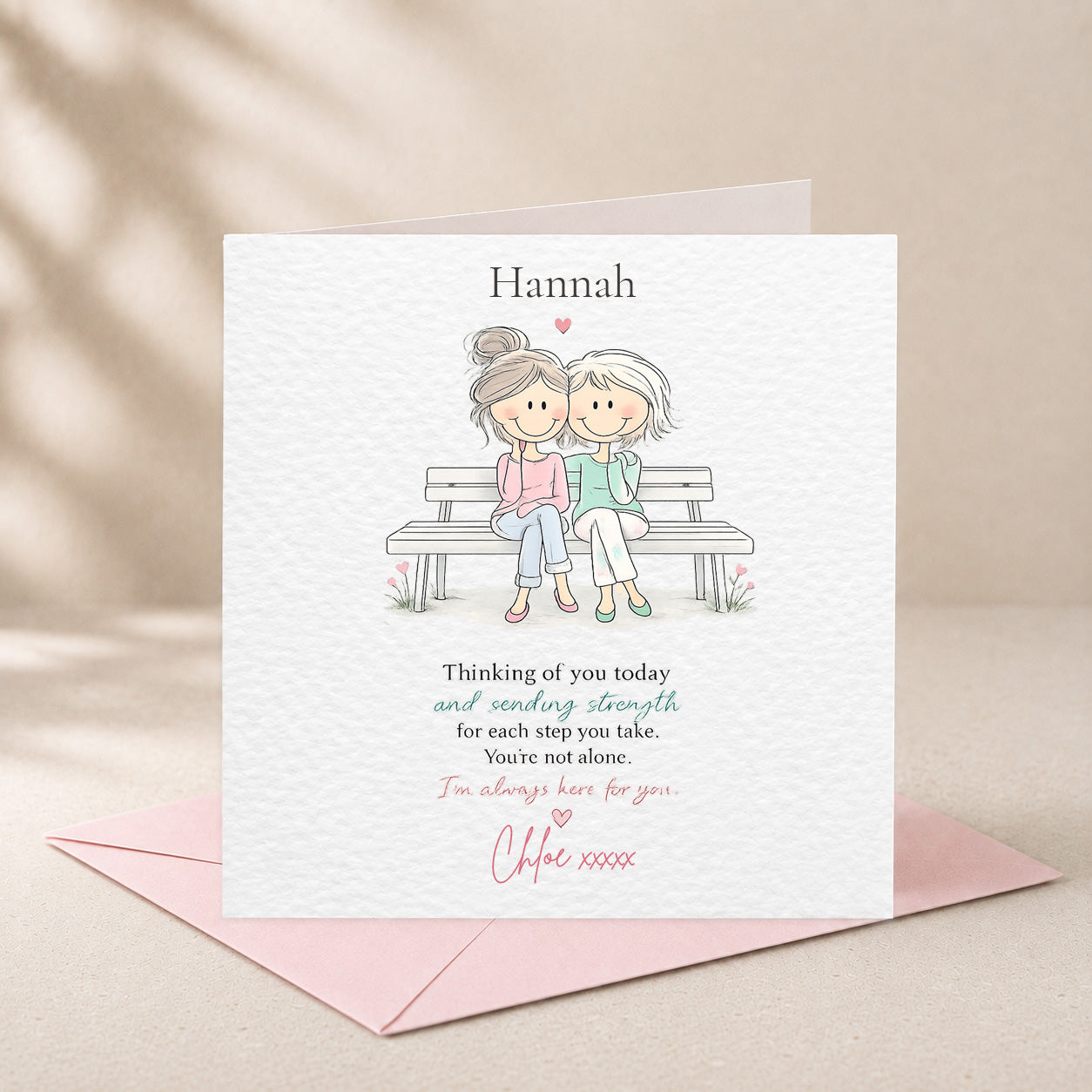 A personalized greeting card with a pink envelope, featuring a watercolor-style illustration of two women sitting on a bench and a sentimental message, displayed on a neutral beige background.