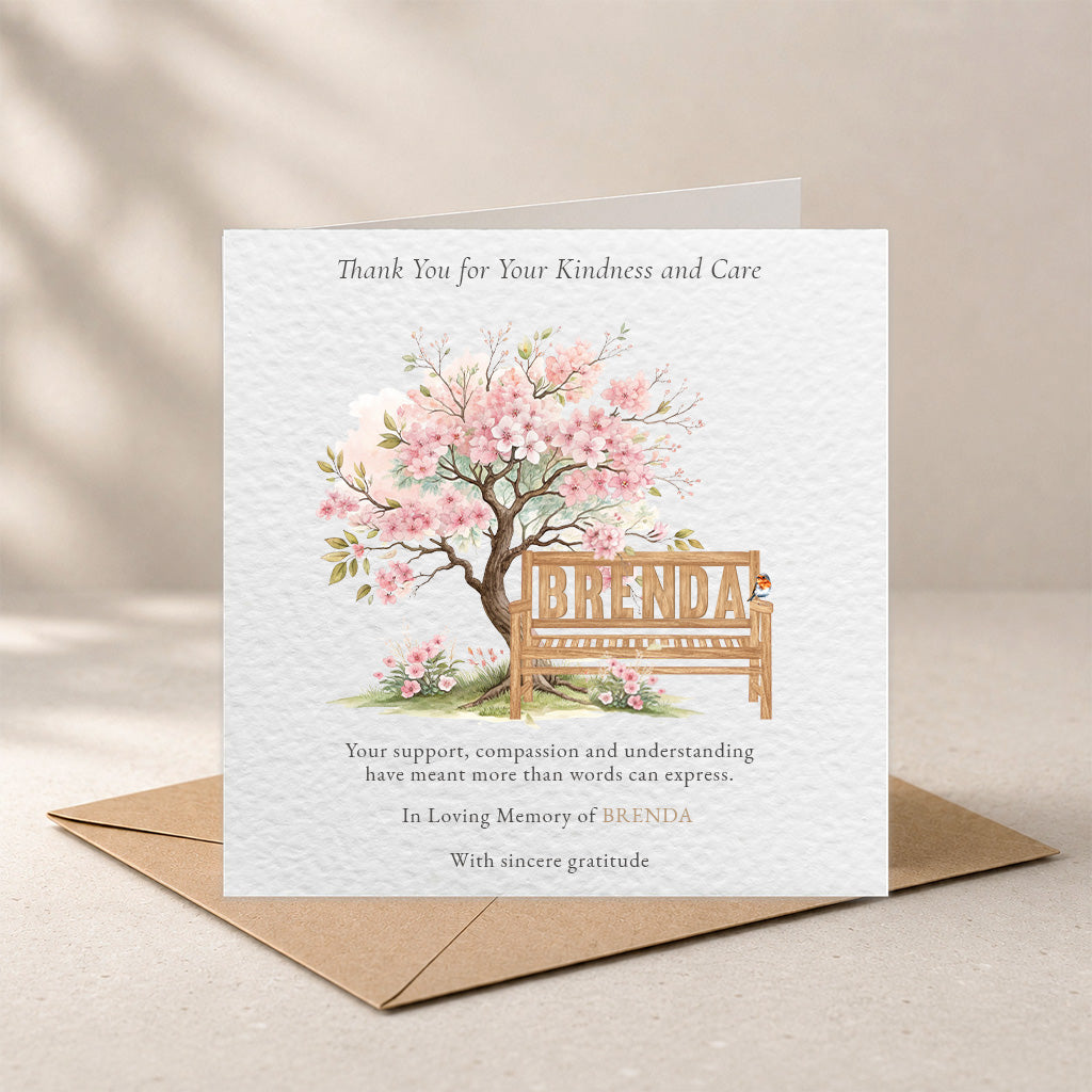 A square white memorial card with a watercolor-style pink flowering tree and wooden bench reading “BRENDA,” shown standing on top of a brown envelope against a soft beige background.