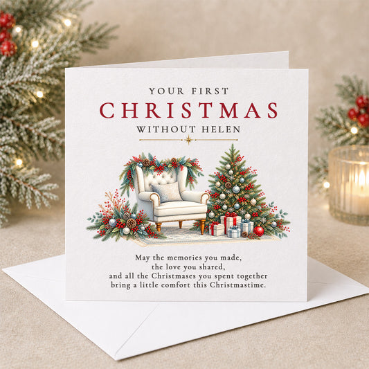 A square Christmas greeting card with an envelope, shown on a beige surface with blurred holiday greenery and lights in the background. The card reads 'YOUR FIRST CHRISTMAS WITHOUT HELEN' above an illustration of a chair, Christmas tree, and gifts, with a memorial-style message below.